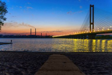 Dnipro Nehri manzarasında bir köprü, duman yayan endüstriyel bacalar ve gün batımında bir balıkçının silueti var. Doğa, endüstri ve insan aktivitelerinin dinamik etkileşimi.