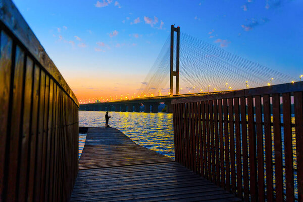 View of a wooden pier leading to a fisherman, set against the backdrop of a cable-stayed bridge over the river at sunset.