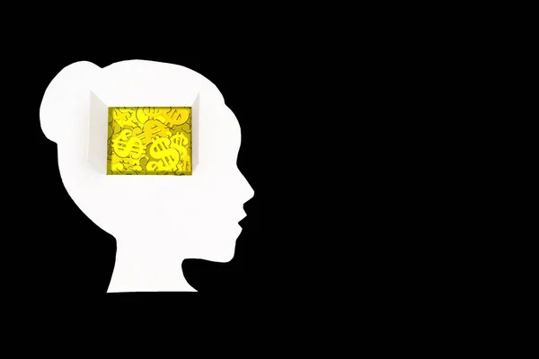 Pile of gold dollar signs seen through the open window in the brain area of a paper woman head cut-out placed on black background with copy space.