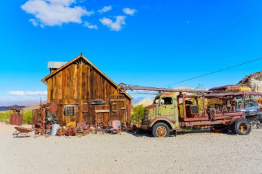 Nelson, Nevada - 15 Nisan 2024: Terk edilmiş eski bir vintage kaldırma kamyonu eski bir ahşap atölyenin yanında, açık mavi gökyüzü altında paslanmış araç ve aksesuarlarla park edilmiş.