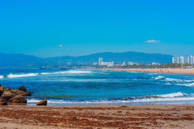 Los Angeles 'ta kumsalda, ön planda kayalar ve deniz yosunları, ortada okyanus, birkaç bina ve arka planda bir dağ silsilesi olan bir şehrin panoramik manzarası..