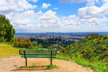 Panoramic view from Runyon Canyon, featuring a solitary green bench on a dirt trail overlooking the expansive landscape of lush greenery and the distant skyline of downtown Los Angeles clipart