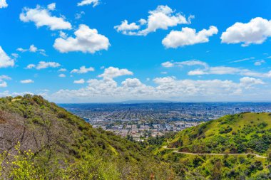 A panorama of Los Angeles under a blue sky with white clouds, and the hills of Runyon Canyon Park with a trail. clipart