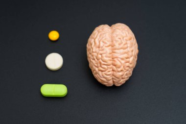 Koyu arka planda yeşil, beyaz ve sarı hapların yanında anatomik beyin modeli, beyin sağlığı ve tedaviyi temsil ediyor.