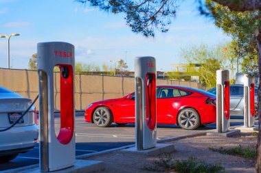 Los Angeles, Kaliforniya - 15 Nisan 2024: Tesla Supercharger istasyonunda ağaçlar ve çitlerle çevrili diğer arabalarla birlikte kırmızı Tesla elektrikli araç şarj ediliyor.