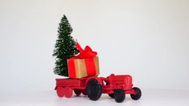 İçinde kırmızı kurdele ve fiyonk olan bir hediye kutusunun olduğu bir traktörün dönüşümlü videosu. Karavanda Noel ağacı taşıyor. Tatil temalı vitrinler için mükemmel..