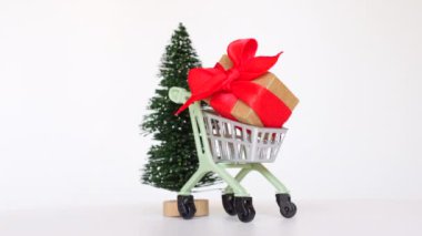 360 derecelik videoda gri arka planda küçük bir Noel ağacı setinin yanında küçük bir alışveriş arabası ve hediye kutusu görülüyor..