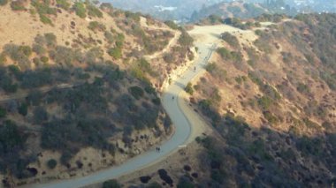 Runyon Canyon Parkı 'ndaki dolambaçlı patikanın hava perspektifinde manzaralı tepelerin arasında yürüyen insanlar var..