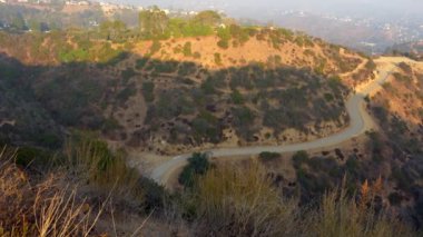 Dolambaçlı bir yolun panoramik manzarası ve Runyon Canyon Parkı 'ndan gelen puslu şehir manzarası doğal manzarayı ve şehir ortamını gözler önüne seriyor..