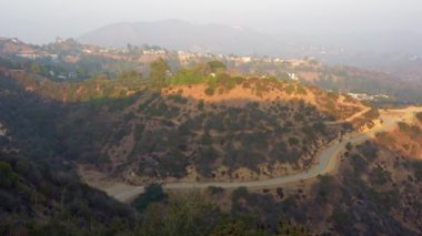 Los Angeles, California - 5 Aralık 2024 Runyon Canyon Parkı 'ndaki tepelerden geçen, doğal manzaraları ve uzak konutları yakalayan manzaralı bir yol görüntüsü.