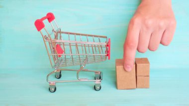 El, üç farklı boyutta minyatür karton kutuyu bir alışveriş arabasına aktarıyor. Bu da teslimat ve e-ticaret konseptini gösteriyor..
