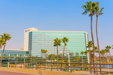 Long Beach, California - 11 Ocak 2025: Modern Hyatt Regency Hotel 'de geniş bir açıdan çekilmiş cam mimari, kaldırım boyunca uzanan palmiye ağaçları ve Long Beach, Kaliforniya' da açık mavi bir gökyüzü bulunmaktadır..