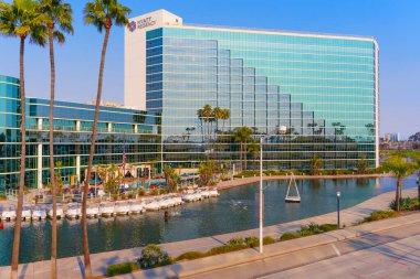 Long Beach, California - 11 Ocak 2025: Long Beach, Kaliforniya 'daki Hyatt Regency Hotel' in çarpıcı manzarası, modern cam cephe, palmiye ağaçları ve altın saat boyunca tekneli sakin bir kanal.