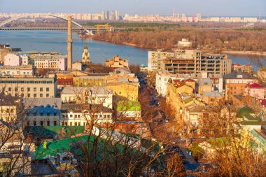 Kyiv, Ukrayna - 9 Mart 2025: Kontraktova Meydanı yakınlarındaki Podil ilçesinin panoramik manzarası, tarihi binalar, nehir manzarası ve sıcak ışık altında yıkanan kentsel manzaranın canlı bir karışımı.