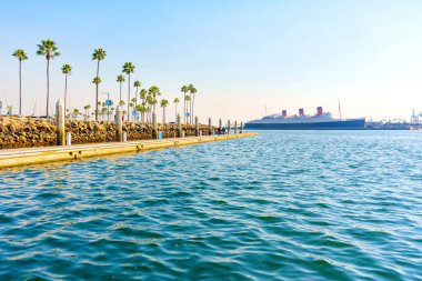 Long Beach, California - 11 Ocak 2025 Long Beach 'in sakin sularından tarihi Queen Mary gemisinin çarpıcı manzarası, palmiye ağaçları ve açık mavi gökyüzü tarafından çerçevelenmiştir..