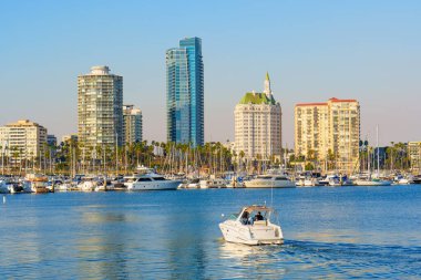 Long Beach, California - 11 Ocak 2025: Yat, Long Beach marinasının sakin sularında süzülüyor, modern yüksek binalarla çevrili, ve altın günbatımı renklerinde yelkenliler, geniş bir açıdan çekiliyor..
