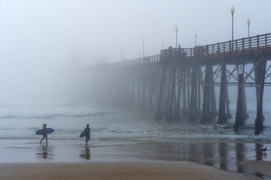 Oceanside, California - 3 Ocak 2025: İki sörfçü, kumlu sahil boyunca Oceanside Belediye İskelesi 'ne doğru yürüyor, sisli ve yumuşak aydınlatmayla kaplı, sakin bir sahil atmosferini yakalıyorlar..