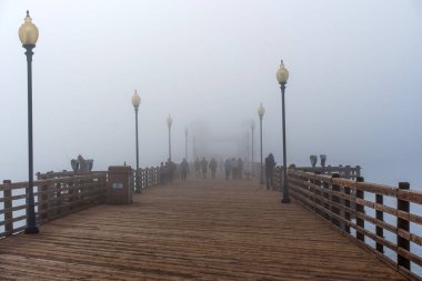 Oceanside, California - 3 Ocak 2025: Oceanside Belediye İskelesi 'nin puslu sabah manzarası balıkçılar ve ziyaretçiler sis kaplı manzaranın keyfini çıkarırken, ahşap yapıyı ve sokak lambalarını vurguluyorlar..