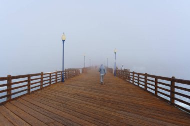 Oceanside, California - 3 Ocak 2025: Okyanus kıyısındaki sisli bir rıhtımda ahşap korkuluklar ve lambalarla çevrili insanlar okyanus kıyısındaki sakin bir atmosferi yakalıyorlar..