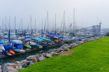 Oceanside, California - 3 Ocak 2025 Oceanside Marina 'da sisli bir sabahın sakin manzarası, arka planda yelkenli tekneleriyle rıhtım boyunca dizilmiş renkli kanolar..