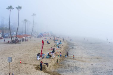 Oceanside, California - 3 Ocak 2025: Aileler, arka planda çocuklar ve renkli plaj ekipmanları ile Oceanside City Beach 'te sisli bir günün tadını çıkarıyorlar..