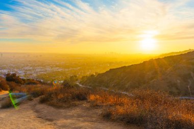 Runyon Canyon Parkı 'ndaki dolambaçlı toprak yolda yürüyen yürüyüşçülerin geniş açılı görüntüsü, arka planda Los Angeles gökyüzünü aydınlatan güzel bir günbatımı var..