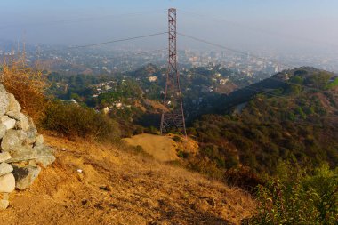Geniş açılı çekim, Runyon Canyon Parkı 'ndan Los Angeles' ın manzaralı manzarasını yakalıyor, engebeli bir arazi ve sisli bir ufuk çizgisine karşı göze çarpan bir güç kulesi var..