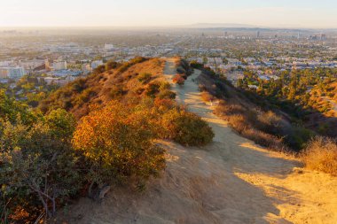 Los Angeles, Kaliforniya - 6 Ocak 2025 Runyon Canyon Parkı 'ndaki kumlu yürüyüş yolunun geniş açılı görüntüsü, Los Angeles ufkunun üzerinde günbatımının canlı renklerini vurguluyor..