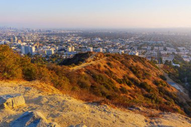 Los Angeles, Kaliforniya - 28 Kasım 2024: Runyon tepelerine bakan toprak patikalardan ve Los Angeles şehir merkezinden altın sise doğru gün batımı manzarası.