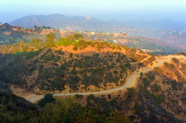 Los Angeles, California - 5 Aralık 2024: Dolambaçlı bir yürüyüş yolunun geniş açılı manzarası lüks yamaçlardaki evleri gösteriyor ve günbatımında Hollywood tabelası görünüyor..