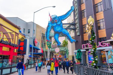 Los Angeles, Kaliforniya - 24 Aralık 2024: Los Angeles 'taki Universal CityWalk' da büyük mavi goril heykeli ve bulutlu gökyüzü altında renkli mağazalar bulunan kalabalık.