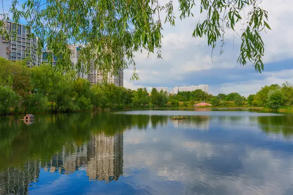 Kyiv 'deki sakin göl, gün boyunca parçalı bulutlu bir gökyüzü altında yeşil ağaçları ve modern mimariyi yansıtıyor..