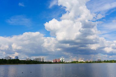 Kyiv ufkunun geniş açılı görüntüsü sakin bir göl boyunca uzanır. Parlak gün ışığında parçalı bulutlu bir gökyüzünün altında binalar sergilenir..