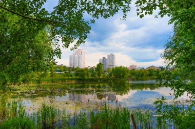 Bereketli yeşillik ve modern yerleşim binalarıyla çevrili sakin bir göl manzarası öğleden sonra saatlerinde parçalı bulutlu bir gökyüzünün altında..