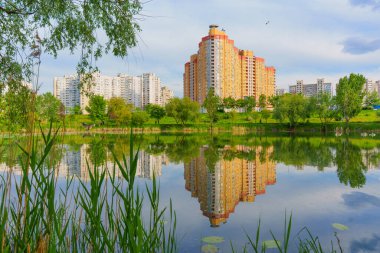 Parçalı bulutlu bir gökyüzünün altında yeşilliklerle çevrili sakin bir göle yansıyan modern yerleşim binalarının geniş açılı görüntüsü.