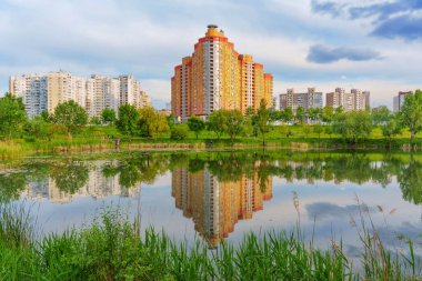 Modern yüksek binalar bulutlu bir günde yeşilliklerle çevrili sakin bir göle yansıyor..