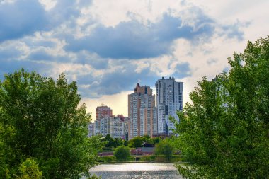 Kyiv, Ukrayna - 19 Mayıs 2025: Kiev 'deki Poznyaky Gölü' nün yanındaki Poznyaky yerleşim bölgesinin yemyeşil ve bulutlu gökyüzü altındaki modern yüksek binaların manzarası.