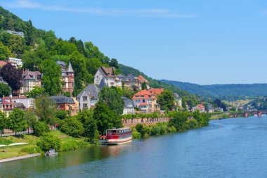 Heidelberg 'deki Neckar Nehri' nin geniş açılı görüntüsü, parlak mavi gökyüzünün altında su kenarında renkli evler ve tekneler yer alıyor..