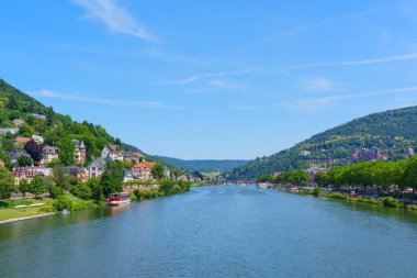 Heidelberg, Almanya 'daki Neckar Nehri manzaralı, açık mavi gökyüzü altında yemyeşil tepeler ve renkli nehir kenarlı evler sergileniyor..