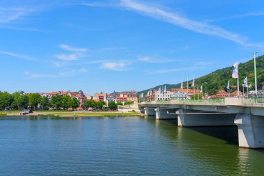Heidelberg, Almanya - 21 Haziran 2025: Heidelberg, Almanya 'daki nehir kıyısının geniş açılı görüntüsü, açık mavi gökyüzü altında bir köprü ve yerleşim binaları.