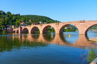 İkonik Eski Köprü Heidelberg 'deki Neckar Nehri boyunca uzanır ve berrak mavi gökyüzünün altında tarihi mimarisini ve dingin su yansımalarını sergiler..