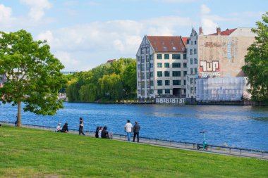 Berlin, Almanya - 30 Mayıs 2025: Berlin 'de yeşil çimenler, modern binalar ve barışçıl bir atmosferle güneşli bir hafta sonunun tadını çıkaran insanları canlandıran canlı bir sahne.