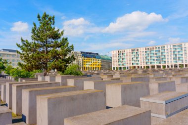 Berlin, Almanya - 31 Mayıs 2025: Berlin 'deki Yahudi Soykırımı Anıtı, büyük beton bloklar, yalnız bir ağaç ve mavi gökyüzü altındaki bulutlu modern binalar.