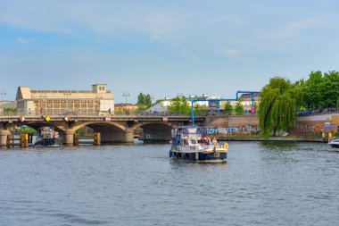 Berlin, Almanya - 31 Mayıs 2025: Berlin, Almanya 'da Spree Nehri üzerinde tekne turu. Arkaplanda açık bir gökyüzünün altında köprülerin, yemyeşil yeşilliğin ve canlı grafitilerin yer aldığı manzara.