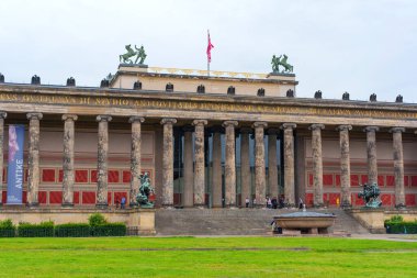 Berlin, Almanya - 1 Haziran 2025: Berlin 'deki Altes Müzesi' nin ön cephesinde bulutlu bir gökyüzüne karşı klasik sütunlar, heykeller ve canlı bayraklar yer alıyor, neoklasik mimariyi vurguluyor.