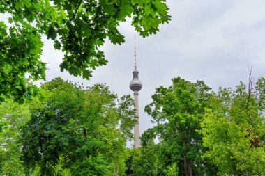 Berlin, Almanya - 1 Haziran 2025: Berlin kentsel park ortamında bulutlu bir gökyüzü altında canlı yeşil yeşillikler çerçeveli Televizyon Kulesi.