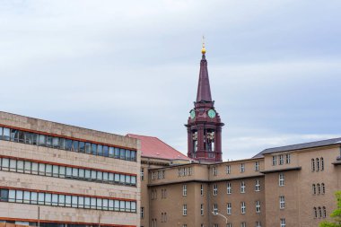 Berlin, Almanya - 1 Haziran 2025: Berlin 'deki çağdaş binalar tarafından çerçevelenmiş saat kulesi, bulutlu gökyüzü altında tarihi ve modern mimarinin bir kesişimini gösteriyor.