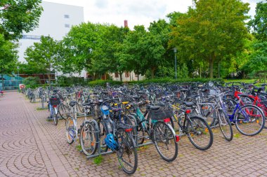 Darmstadt, Almanya - 16 Haziran 2025: Darmstadt, Almanya 'da parçalı bulutlu bir gökyüzünün altında yeşil ağaçların olduğu bir zemin üzerine kurulmuş sayısız bisikletle dolu bisiklet park alanı.