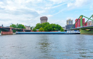 Frankfurt, Almanya - 10 Haziran 2025: Frankfurt 'taki ana nehir boyunca ilerleyen kargo nakliye gemisi, modern köprüler ve kentsel binalar ile canlı bir endüstriyel ortam.