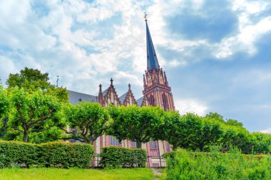 Frankfurt, Almanya - 10 Haziran 2025: Iconic kilise kulesi Frankfurt 'ta bulutlu bir gökyüzüne canlı yeşil ağaçların arasından bakıyor.